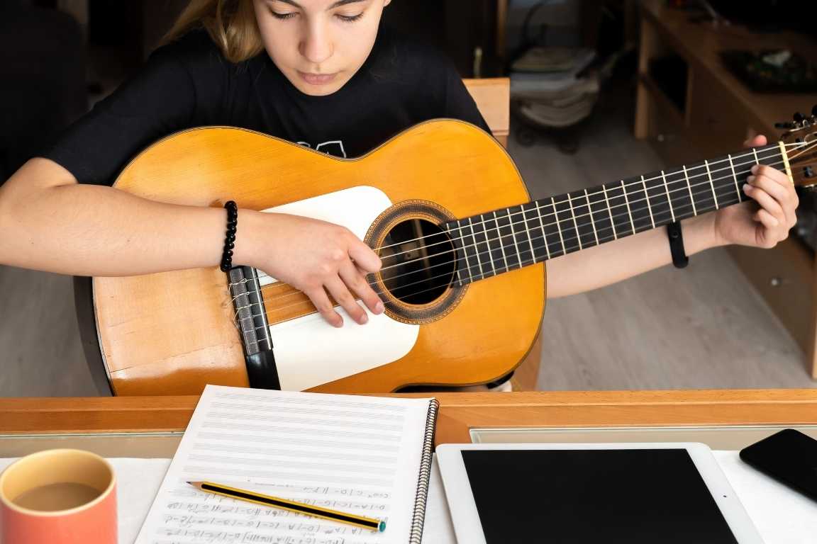 Persona estudiando guitarra con partituras, cuaderno y tableta sobre la mesa